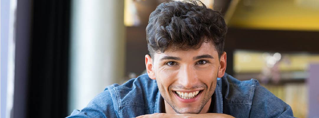 A smiling student rests his chin on his hands over a pile of books