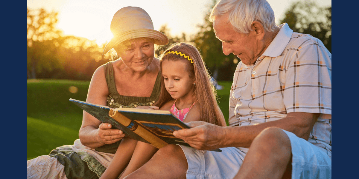 A young girl sits between her grandparents, reading a book together in a sunlit outdoor setting. The grandmother wears a sun hat, while the grandfather smiles at the girl, creating a warm family moment.