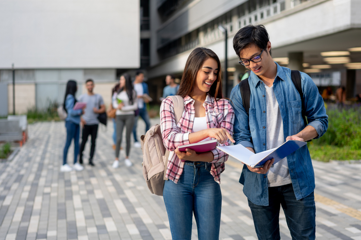 In the foreground, female and male students look at a book the male student is holding. In the background, a group of students visits on a college campus.