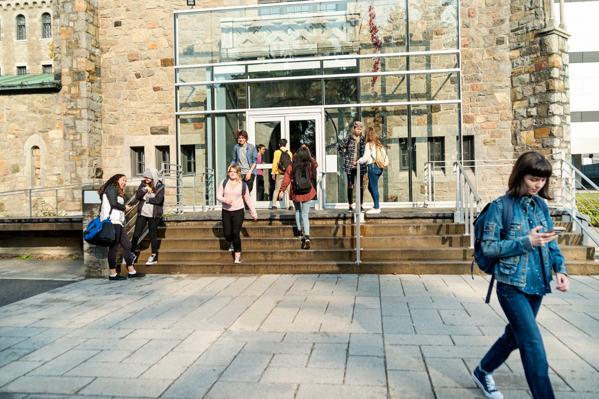 A female student walks away from a group of students gathered on the steps in front of a college building.