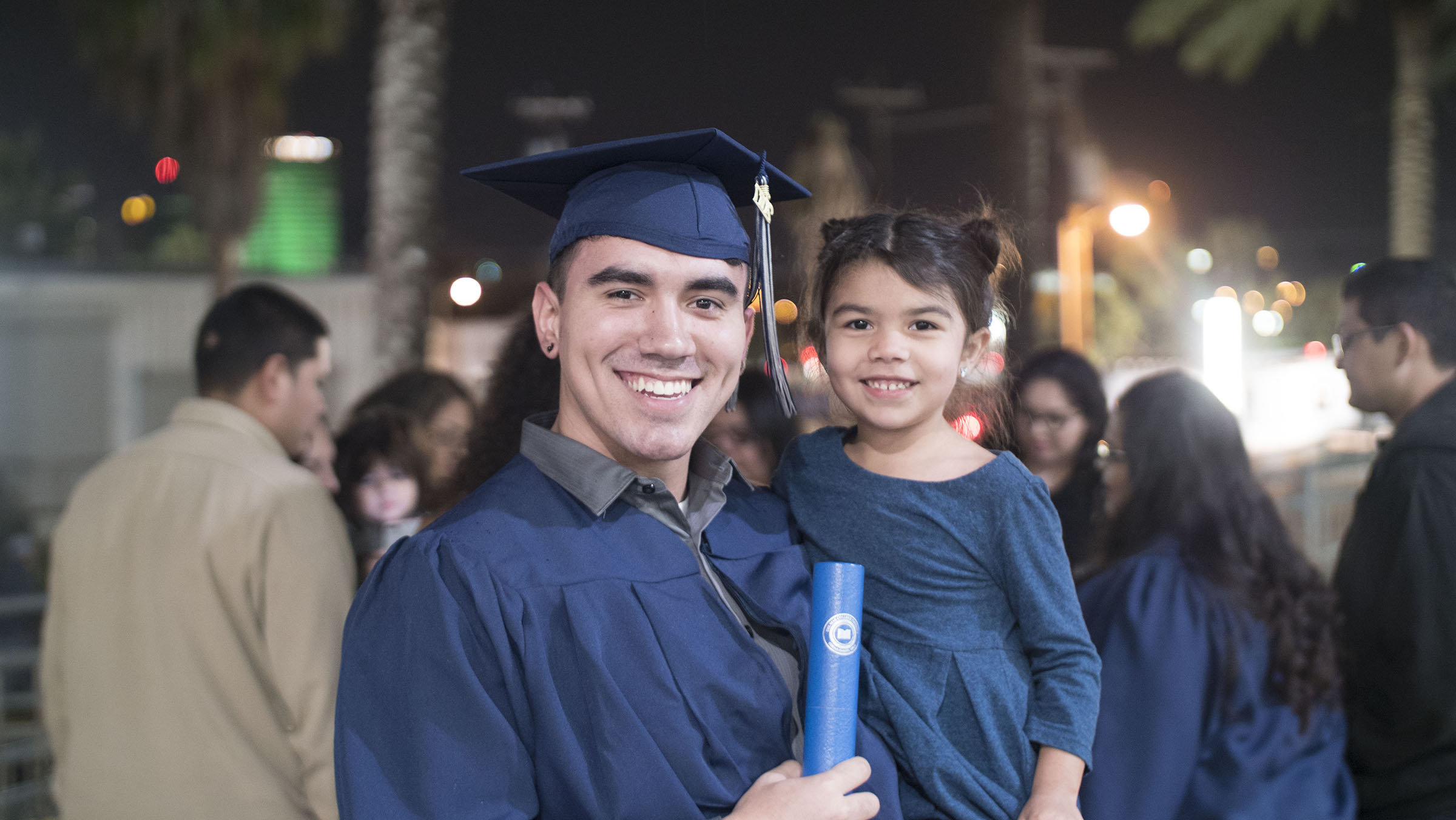 A smiling student poses in cap and gown while holding his degree and a young child