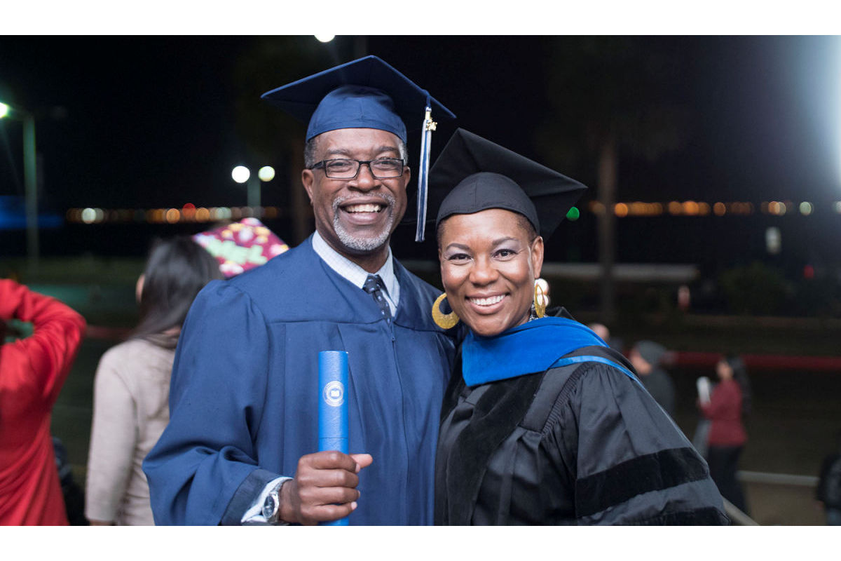 A man and woman in graduation robes smile for the camera