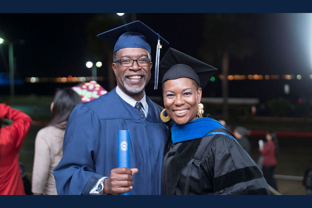 A man and woman in graduation robes smile for the camera