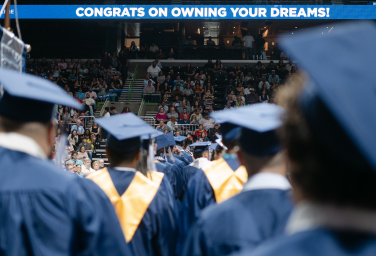 Graduates in blue caps and gowns face a seated crowd during a graduation ceremony beneath a banner.