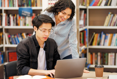An instructor and student work together on a laptop in a library, with bookshelves behind them and a coffee cup on the table.