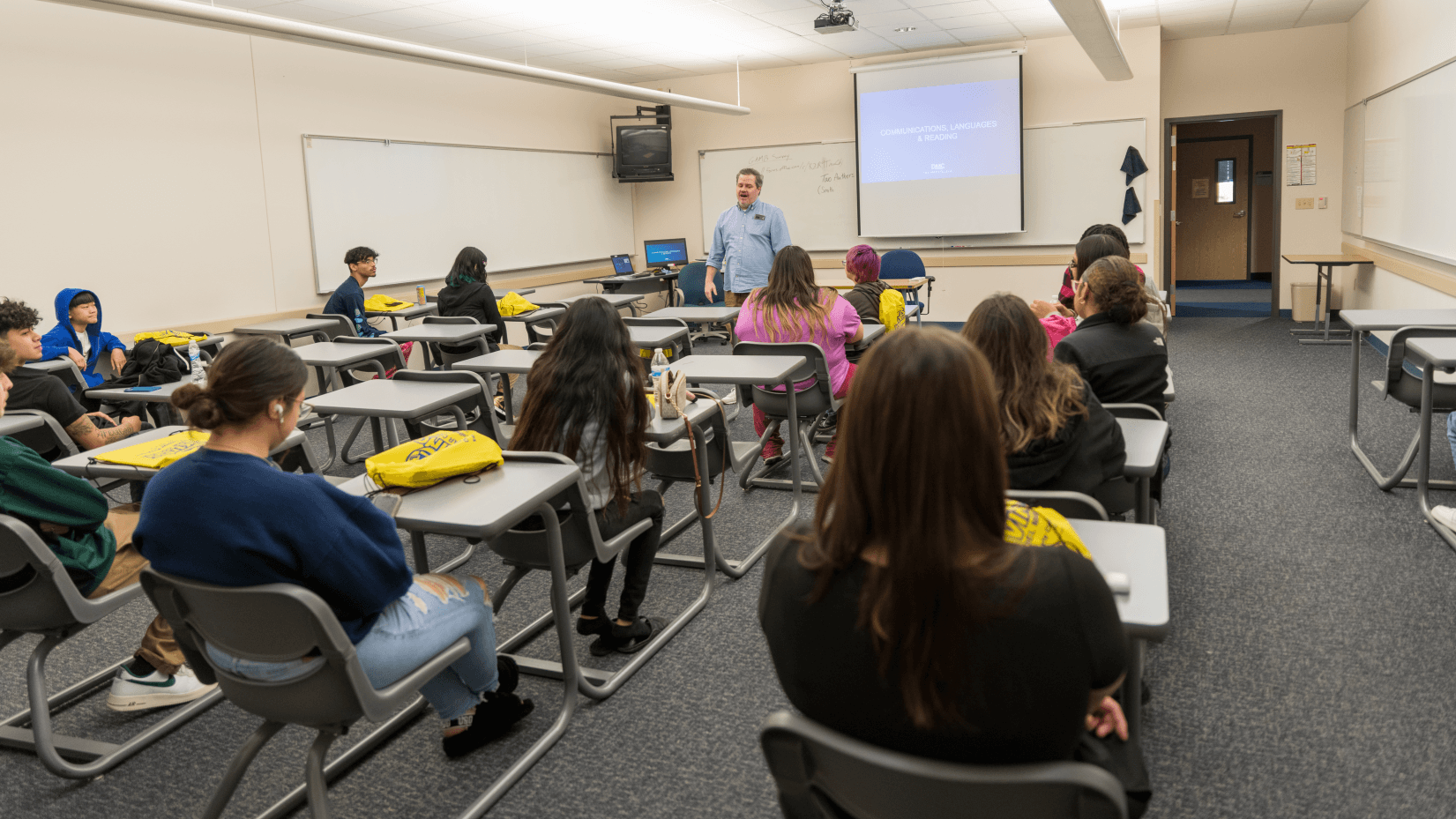 Instructor addressing students in a classroom with a projected presentation screen and whiteboards.