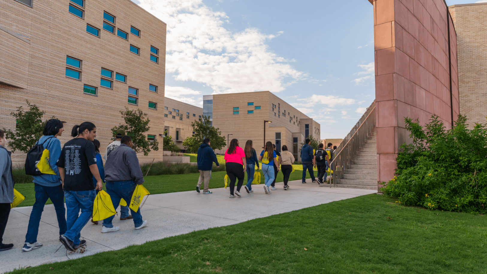 Students carrying yellow bags walk between tan-brick campus buildings on a landscaped pathway.