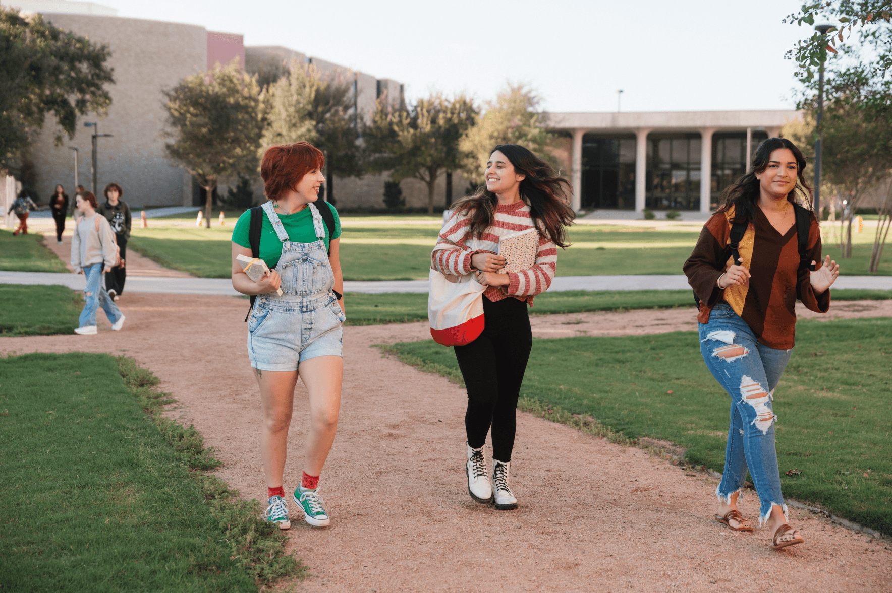 Three students walk along a landscaped campus path, with campus buildings and trees in the background.