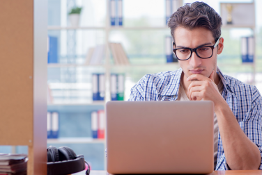 Man in glasses working thoughtfully on a laptop at a desk, with headphones beside him and bookshelves in the background.