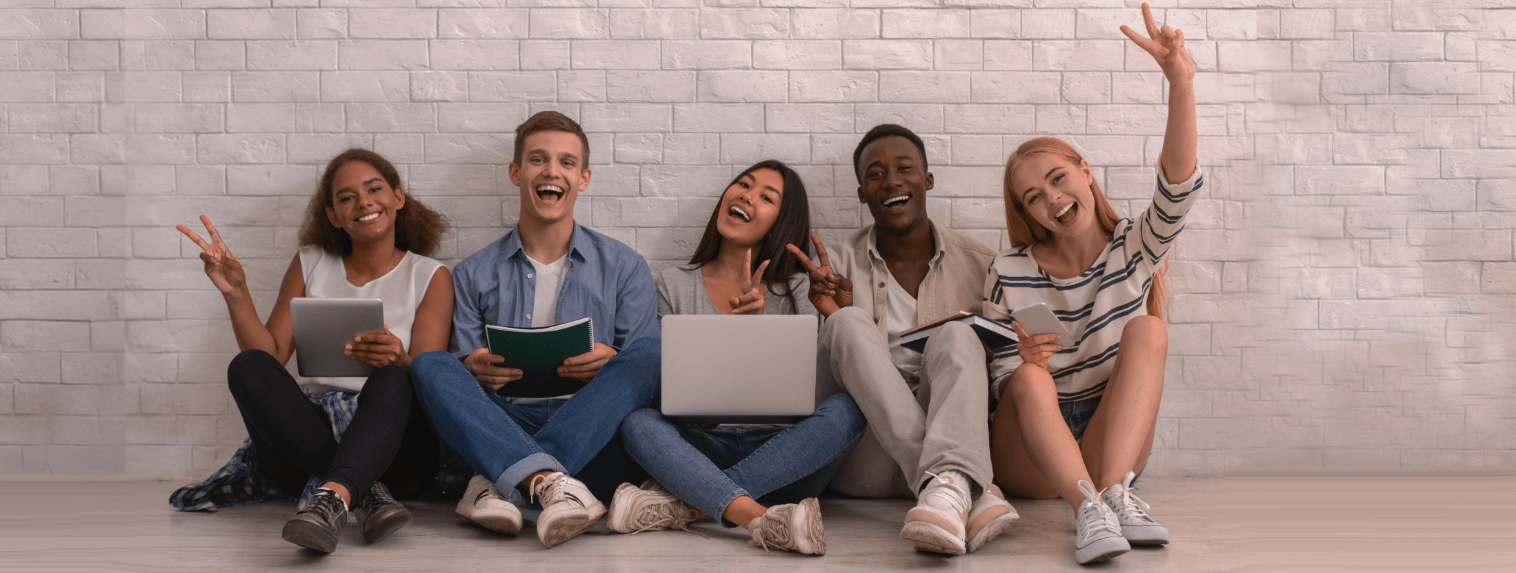Five young adults sitting on the floor against a light brick wall, smiling and holding electronic devices and notebooks.