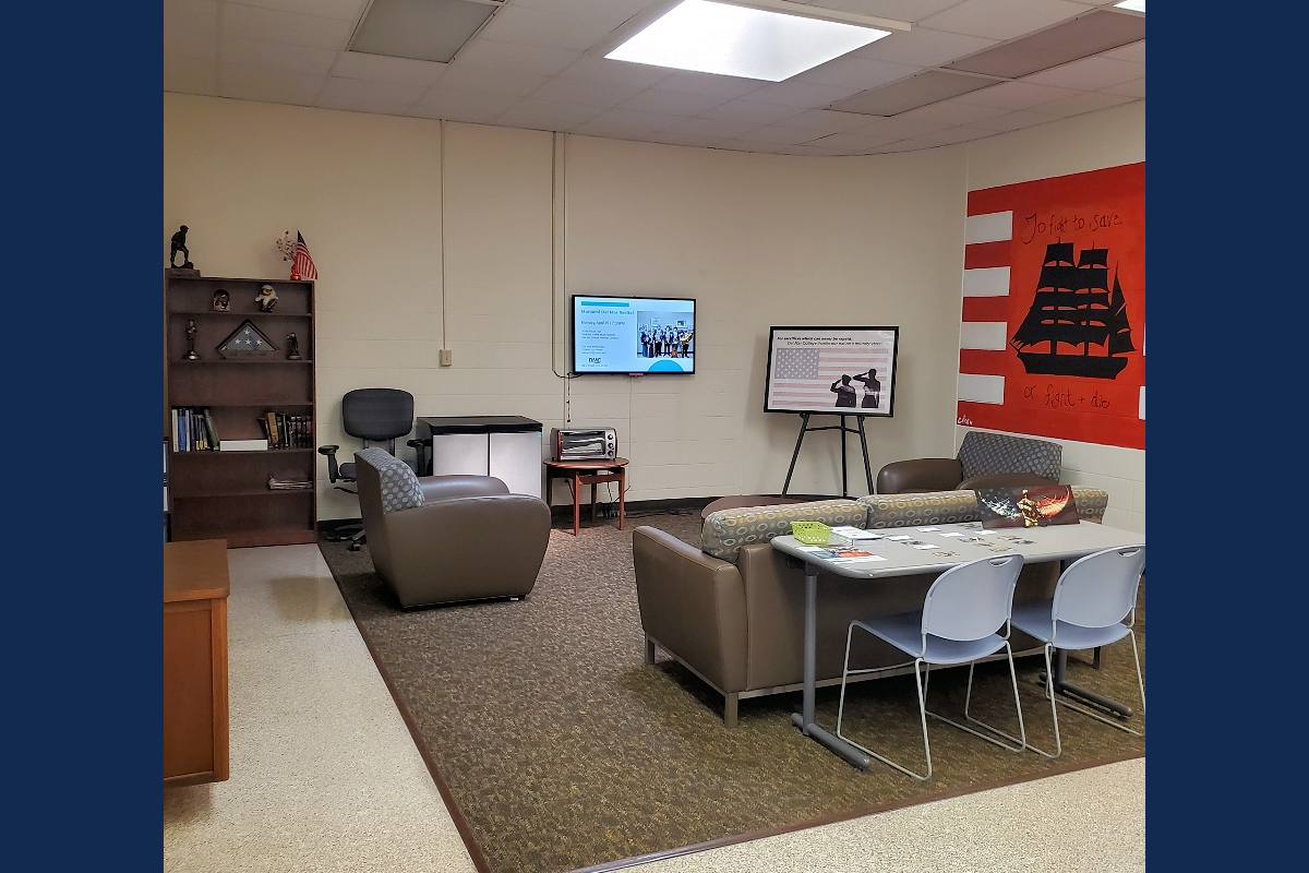 Windward Campus Veteran's lounge with chairs, a couch, bookshelf, and a mounted TV displaying a presentation.